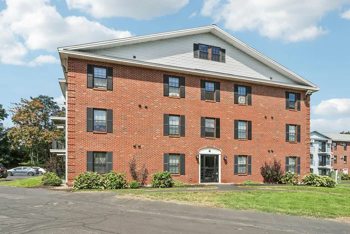 A classic three-story red brick apartment building with white trim, a gabled roof, and beautiful landscaping at our pet-friendly apartments in Worcester, MA. | Princeton Place Apartments