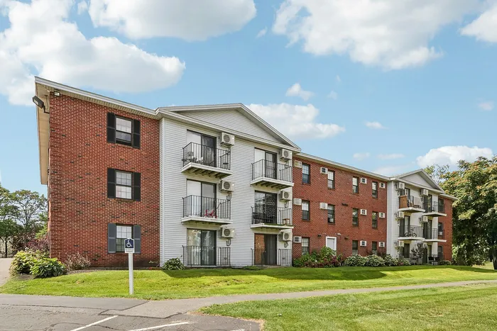 Princeton Place Apartments | Princeton Place Apartments in Worcester, MA is a three-story brick and white-sided building with balconies and window air conditioners, set against a blue sky with clouds, surrounded by green grass and a sidewalk. | Worcester Apartments with Virtual Tours
