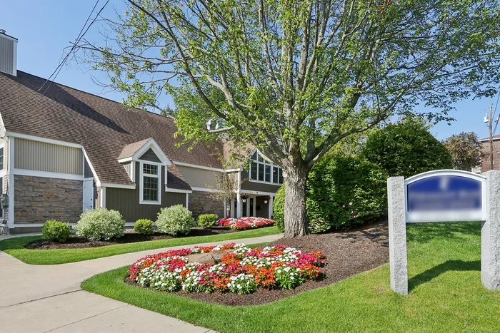 Pheasant Run Apartments | A modern clubhouse with large windows and a brown roof stands behind a swimming pool surrounded by lounge chairs at Pheasant Run Apartments in Nashua, NH, all beneath a clear blue sky. | Apartments with Balcony in Nashua