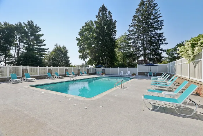 Pheasant Run Apartments | A modern clubhouse with large windows and brown roof stands behind an outdoor swimming pool at Pheasant Run Apartments in Nashua, NH, surrounded by turquoise lounge chairs and a white fence on a sunny day. | Apartments with Air Conditioning in Nashua