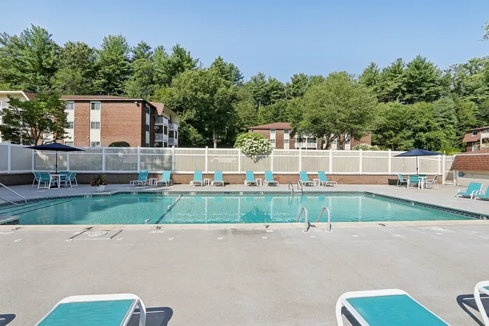Pheasant Run Apartments | Outdoor swimming pool surrounded by lounge chairs and tables at Pheasant Run Apartments, next to the leasing office/clubhouse with large windows and brown roof in Nashua NH, under a clear blue sky. | Nashua Apartments with Pickleball Court