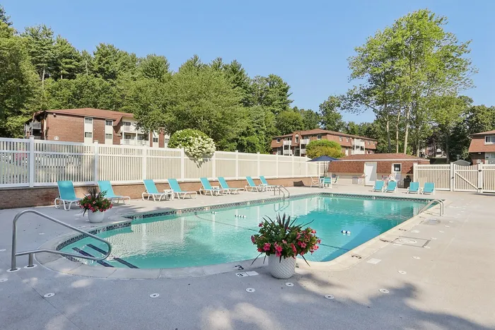 Pheasant Run Apartments | A clear outdoor swimming pool at Pheasant Run Apartments in Nashua, NH, is surrounded by lounge chairs and tables, with a fence and trees in the background next to a building under a sunny sky. | Nashua Apartments with Virtual Tours