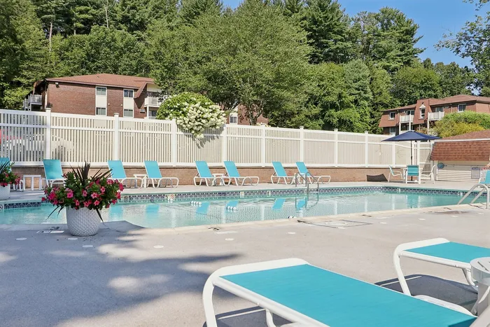 Pheasant Run Apartments | Enjoy the outdoor swimming pool at Pheasant Run Apartments in Nashua NH, featuring turquoise lounge chairs and tables, a white fence, tall green trees, umbrellas, and potted plants under a clear blue sky. | Apartments with Washer & Dryer in Nashua