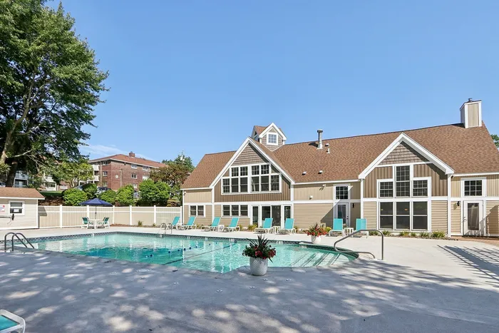Pheasant Run Apartments | Enjoy the bright, sunny days at Pheasant Run Apartments in Nashua NH, featuring an outdoor swimming pool area with turquoise lounge chairs under a white pergola, surrounded by a white fence and lush trees beneath clear blue skies. | Apartments with Balcony in Nashua