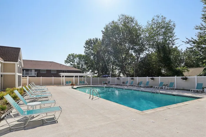 Pheasant Run Apartments | Relax by the outdoor swimming pool at Pheasant Run Apartments in Nashua, NH, featuring turquoise lounge chairs on a concrete deck. A modern building with large windows and a brown roof sits nearby amid trees and a clear blue sky. | Apartments with Washer & Dryer in Nashua