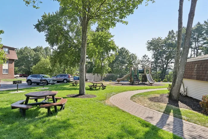 Pheasant Run Apartments | A landscaped building entrance at Pheasant Run Apartments in Nashua NH features colorful flower beds, neatly trimmed shrubs, a large tree, and a blank blue sign on a lawn along a curved sidewalk. | Nashua Apartments with Virtual Tours