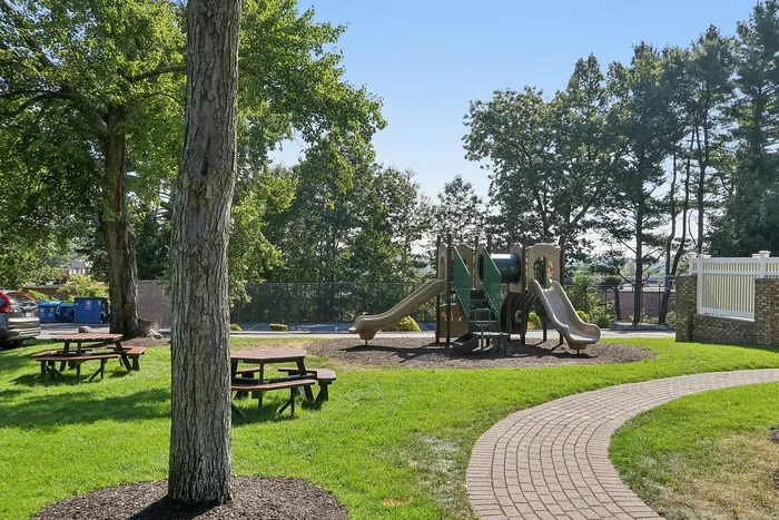 Pheasant Run Apartments | Outdoor swimming pool at Pheasant Run Apartments in Nashua, NH, surrounded by lounge chairs, potted plants, and a white fence, with brick apartment buildings and green trees under a clear blue sky. | Wheelchair Accessible Apartments in Nashua