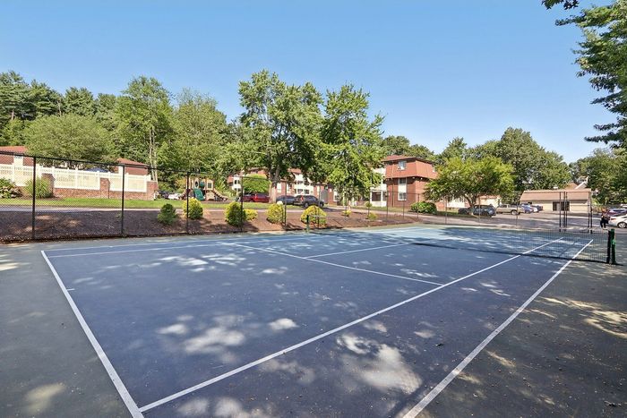Pheasant Run Apartments | Outdoor swimming pool surrounded by blue lounge chairs, white fence, and potted plants at Pheasant Run Apartments, with brick apartment buildings and tall green trees in Nashua NH on a sunny day. | Apartments with Air Conditioning in Nashua