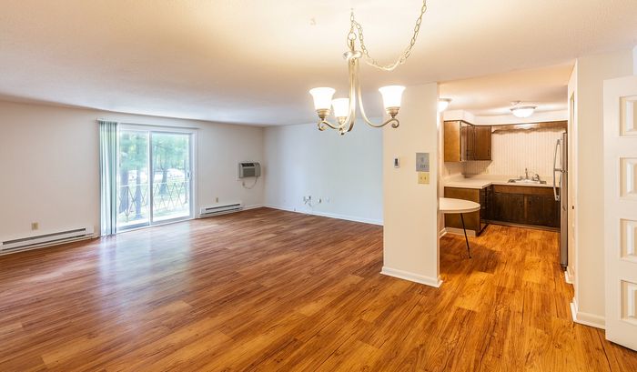 Inviting apartment featuring a spacious open living area with warm wood flooring, elegant chandelier lighting, and sliding glass doors leading to a serene outdoor view.| The Royal Crest | Apartment in North Andover, MA