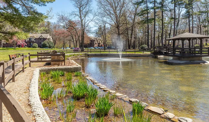 Charming outdoor view of a serene pond with a fountain and a picturesque wooden bridge surrounded by lush greenery and blooming trees.| The Royal Crest | Apartment in North Andover, MA