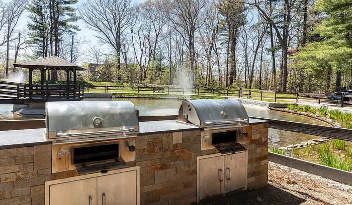 Inviting outdoor space with dual stainless steel grills overlooking a peaceful pond and gazebo, perfect for relaxed gatherings.| The Royal Crest | Apartment in North Andover, MA