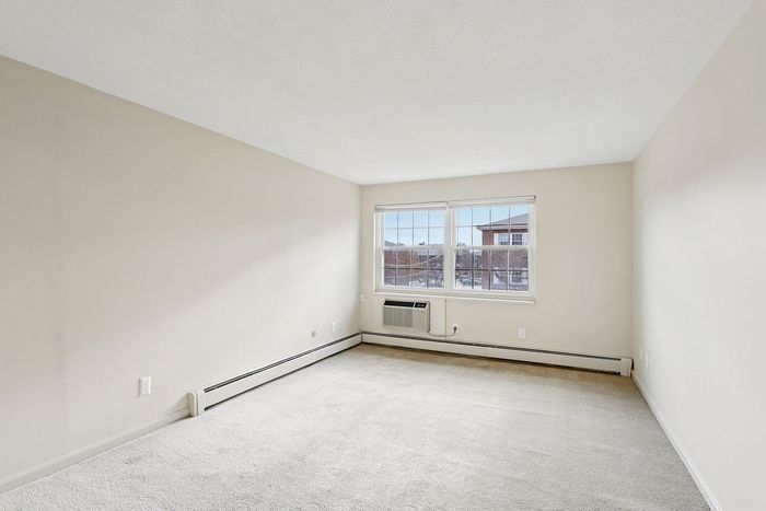 Bright and spacious room with large window and built-in air conditioning unit, featuring fresh neutral-toned walls and plush carpeting.