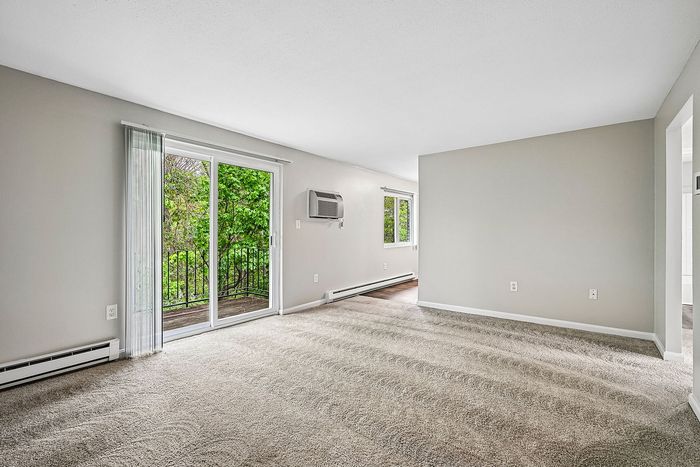Bright and airy living room with plush carpet, large windows, and sliding glass doors opening to a lush green balcony view.