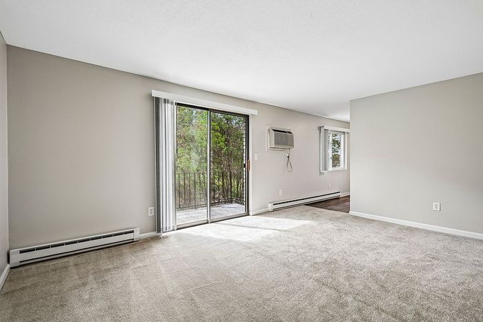 Bright and airy living room with plush carpeting, large sliding glass door opening to a charming balcony, featuring serene outdoor views and abundant natural light.