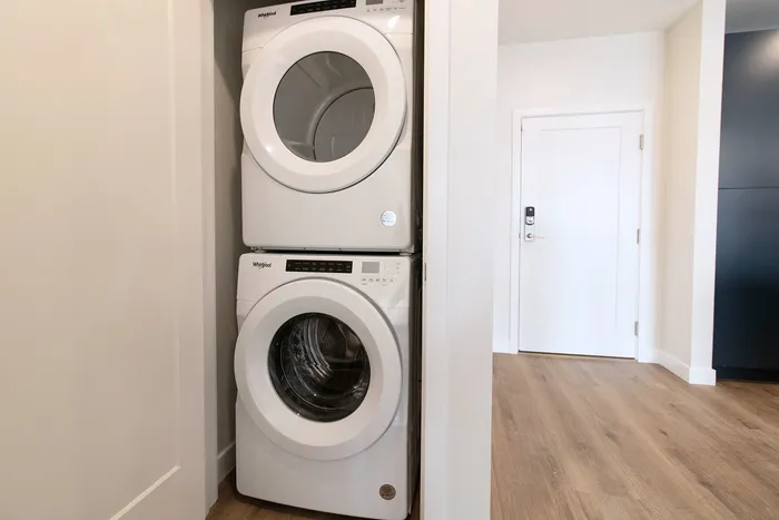 Modern apartment with a sleek, stacked washer and dryer set, featuring elegant wood flooring and a stylish white door, enhancing convenience and elegance.