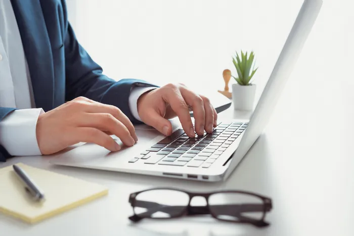 Professional workspace setup with a sophisticated laptop user in a suit, accompanied by a notebook, pen, and stylish eyeglasses, enhanced by a small green plant for a touch of nature.