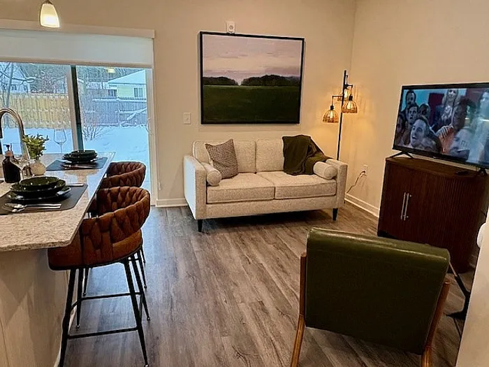 Inviting living area featuring cozy seating, sleek flat-screen TV, and stylish dining counter with elegant bar stools, all bathed in natural light from the sliding door to a charming snowy backyard view.