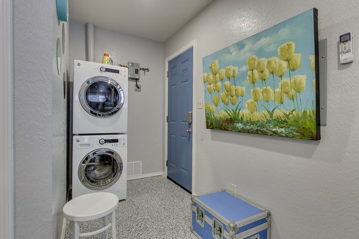 Modern laundry room with stacked washer and dryer, bright blue door, and artistic floral painting.