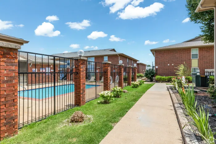 Charming brick apartment complex featuring a refreshing pool, manicured gardens, and a sunny walkway.