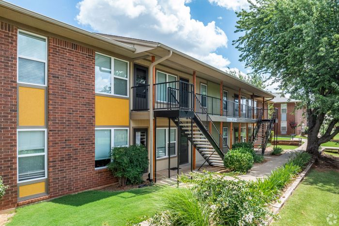 Charming two-story apartment building with vibrant yellow accents, lush green landscaping, and modern exterior staircases, set against a clear blue sky.