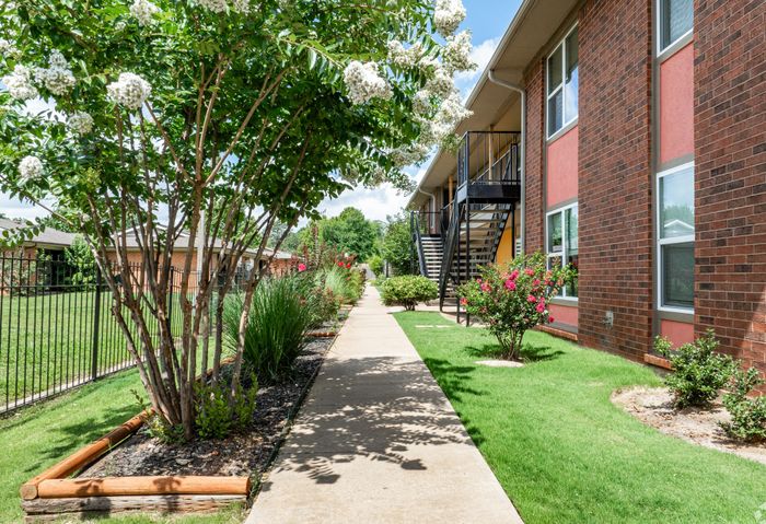 Charming apartment exterior with vibrant landscaping, featuring lush greenery, colorful flowers, and a brick building under a clear sky.