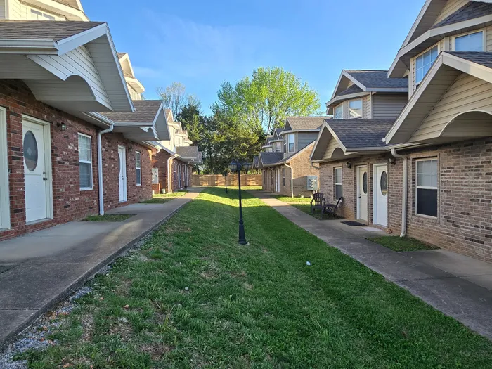 Charming apartment complex with red brick exteriors, nestled along a lush green pathway, offering cozy and connected living spaces.