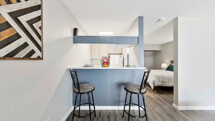 Modern apartment interior highlighting a cozy breakfast bar with two stools, next to a sleek kitchen area and a glimpse of a stylish bedroom.