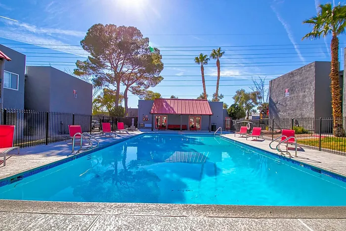 Inviting outdoor pool with red lounge chairs surrounded by modern apartments and palm trees under a bright blue sky.