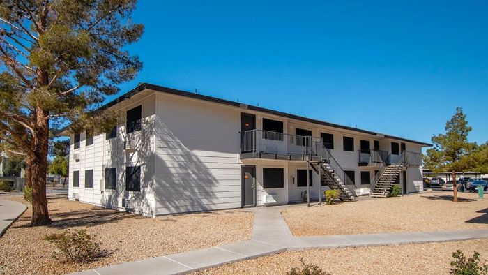Charming two-story apartment building surrounded by scenic landscaping and vibrant trees, offering a peaceful living environment with accessible stairs and pathways under a clear blue sky.