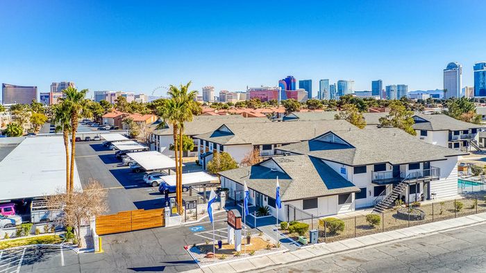 Modern apartment complex with palm trees and ample parking, set against a vibrant cityscape with high-rise buildings and a clear blue sky.