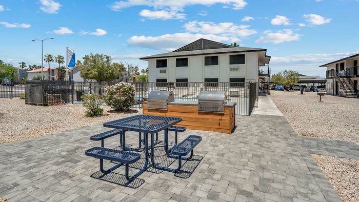 Inviting outdoor communal area with modern grills, stylish picnic seating, and a charming apartment complex backdrop set under a beautiful blue sky.