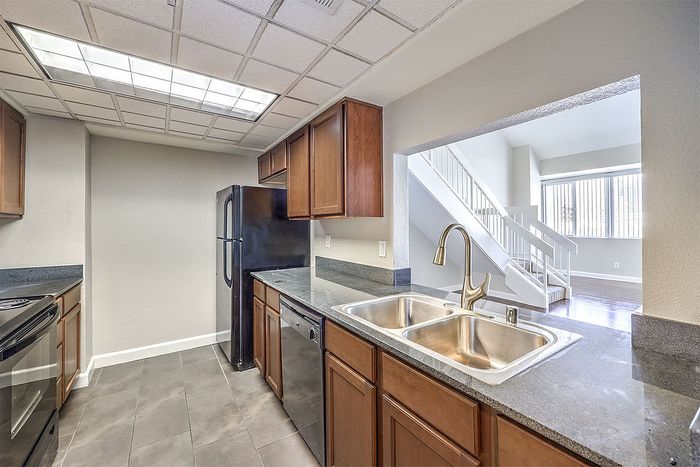 Modern kitchen with sleek dark countertops, stainless steel appliances, and ample cabinetry adjacent to a bright, open living area with large windows and stylish stairs.