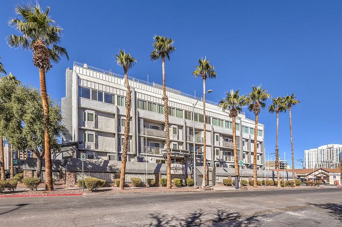 Modern urban apartment building with sleek design, towering palm trees in front, set against a clear blue sky.