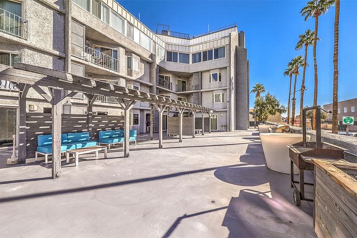 Modern apartment exterior with spacious patio, stylish pergola, and vibrant seating area under clear blue skies with palm trees.