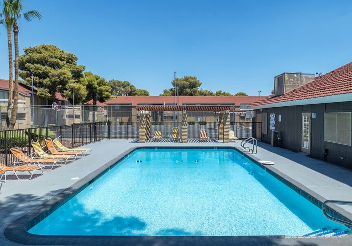 Inviting outdoor pool area with sun loungers, surrounded by lush greenery and modern apartment buildings under a clear blue sky.