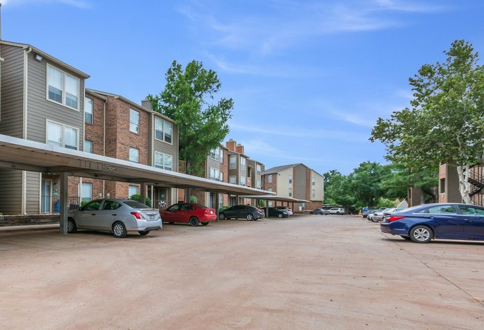 Charming apartment complex with modern designs, ample parking, and lush greenery, under a bright blue sky.