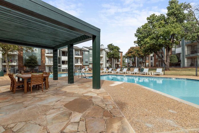 Modern apartment complex with inviting outdoor pool and stylish seating area under a pergola, surrounded by lush landscaping.