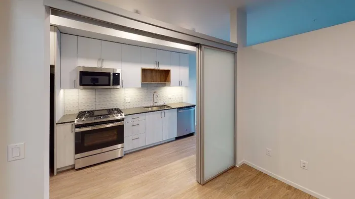 View from den looking into galley-style kitchen with stainless steel appliances in apartment at Modera River Train in Redmond, WA.