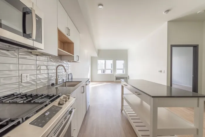 Closeup of kitchen with moveable kitchen island with storage beneath, gas range, gray countertops, white tile backsplash and stainless steel appliances with living room in background