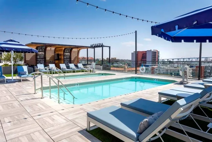 Rooftop pool with chaise lounge seating and umbrellas under string lighting looking out over the city and bay at Modera San Diego apartments.