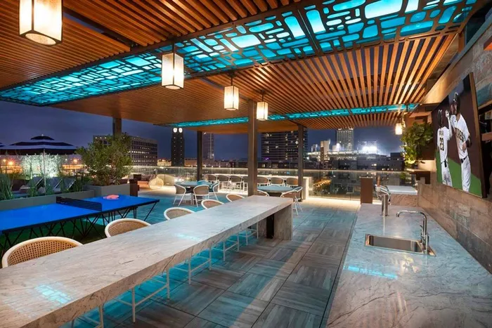 Rooftop lounge area at night looking from outdoor kitchen onto gaming area and pool deck with a Padres game on the TV at Modera San Diego apartments.