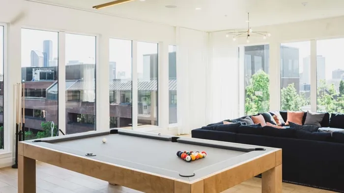 Light-filled room surrounded by large windows looking out on the city with lounge seating and a pool table