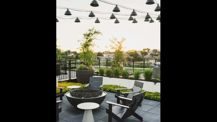 Adirondack chairs surround a round fire put under string lighting on the rooftop surrounded by greenery at Modera Broadway apartments.