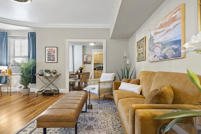 Chic living room with golden hues, modern decor, and open access to a cozy dining area. Inviting natural light enhances the elegant furnishings and artwork.