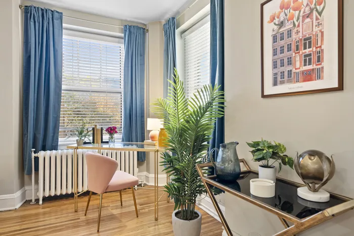 Charming home office nook featuring a pink velvet chair, elegant gold desk, lush green plant, and large windows with blue curtains.
