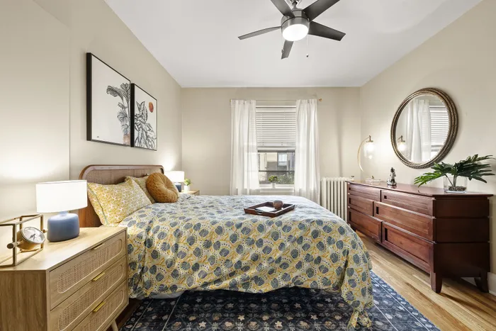 Inviting bedroom with modern ceiling fan, elegant wall art, and plush bedding. Natural light pours through large windows, highlighting a stylish wooden dresser and cozy ambiance.