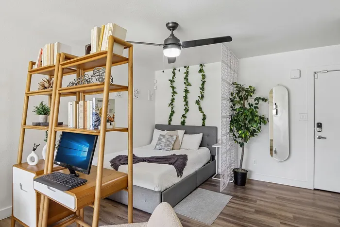 Sleek modern apartment room with cozy bed, stylish ceiling fan, and elegant divider. Features a versatile wooden shelving unit with a built-in workspace, perfect for productivity. Accented by lush greenery for a refreshing touch.