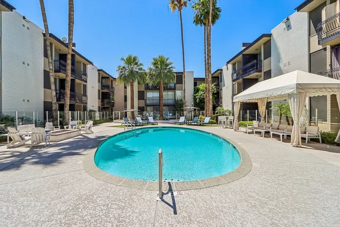 Luxurious apartment complex featuring a sparkling circular pool, surrounded by elegant lounge chairs and tall palm trees.