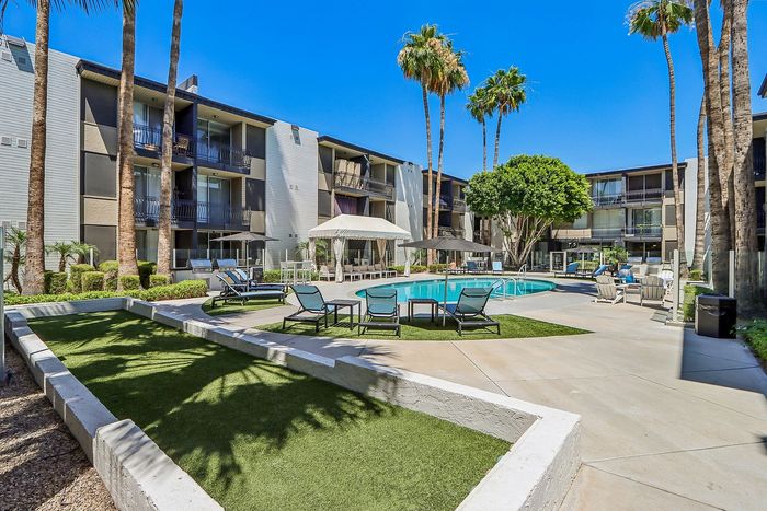 Inviting apartment complex courtyard featuring a sparkling pool, sun loungers, and lush palm trees under a clear blue sky.