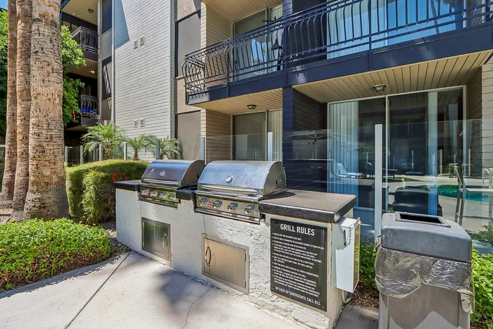 Modern outdoor grill area with stainless steel barbecues, lush greenery, and shaded balconies in a stylish apartment setting.